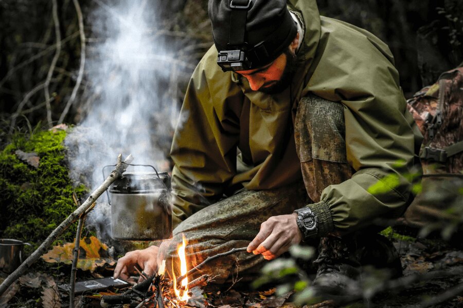 Muž v membránovej bunde pri ohni v lese. Zdroj: https://www.pexels.com/photo/kneeling-man-starting-fire-in-a-fireplace-15538110/