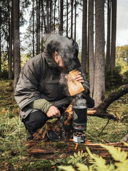 Muž v zateplenej bunde kľačí v lese pri variči Jetboil, z ktorého stúpa para. Pripravuje teplé jedlo alebo nápoj ako zdroj energie a tepla počas jesennej výpravy. Foto: Rigad