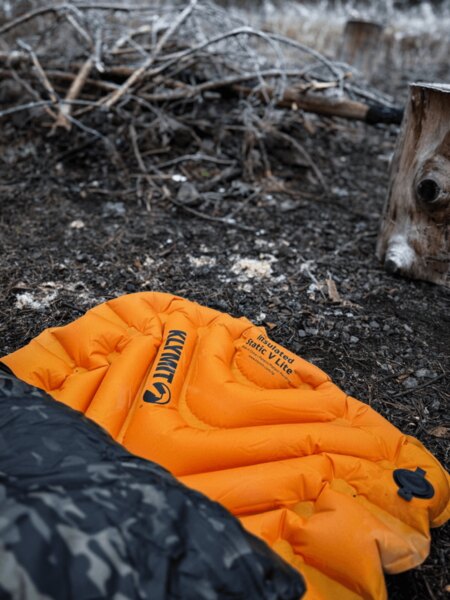 Nafukovacia karimatka Klymit Insulated Static V Lite v oranžovej farbe na zemi pri ohnisku, čiastočne zakrytá spacákom s maskovacím vzorom. Izolácia od chladu a vlhkosti pri spaní v teréne. Foto: Rigad