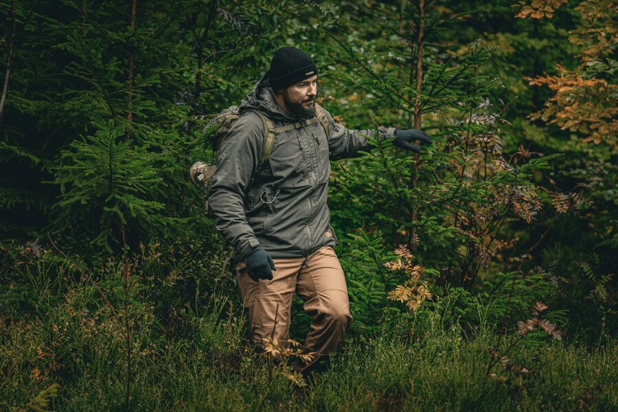 Muž v jesennom lese v sivej zimnej bunde Yukon s izoláciou Climashield™ Apex<sup>®</sup> od Helikon-Tex<sup>®</sup>, čiapke a rukaviciach, s batohom; ukážka správneho vrstvenia a ochrannej vrchnej vrstvy do vlhka a vetra. Foto: Rigad