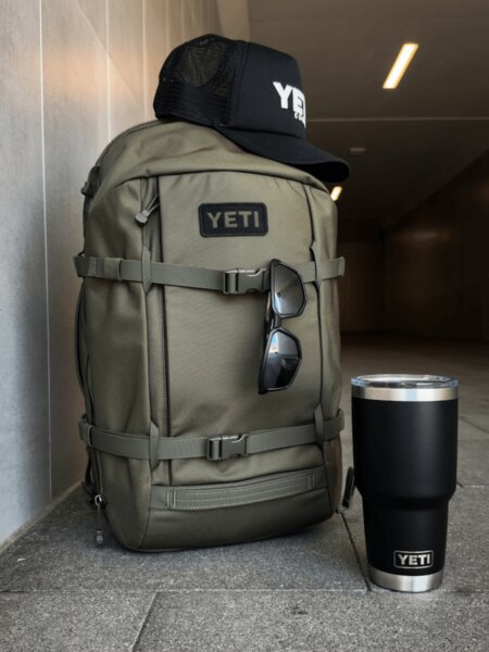 Svetlosivý batoh YETI Ranchero 27 l s čiapkou a čiernou fľašou YETI Rambler.